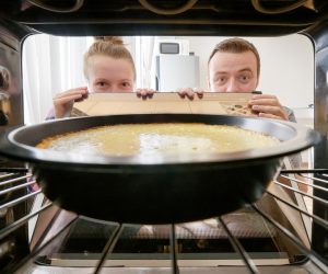 Young woman and young man staring at cheesecake into oven in kitchen. View from inside of the oven. Housewife and her husband holding the oven door.
